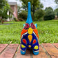 A glossy talavera ceramic cat figurine decorated with vibrant hand-painted patterns in turquoise, yellow, green and red, standing on a brick path with a green lawn and a house in the blurred background. Back tail view.