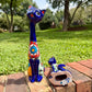 Two colorful hand-painted ceramic cat figurines displayed outdoors on a brick path with a green grassy lawn and trees in the background. One figurine is standing upright and is deep cobalt blue with red, orange, yellow, and white painted patterns on its body.