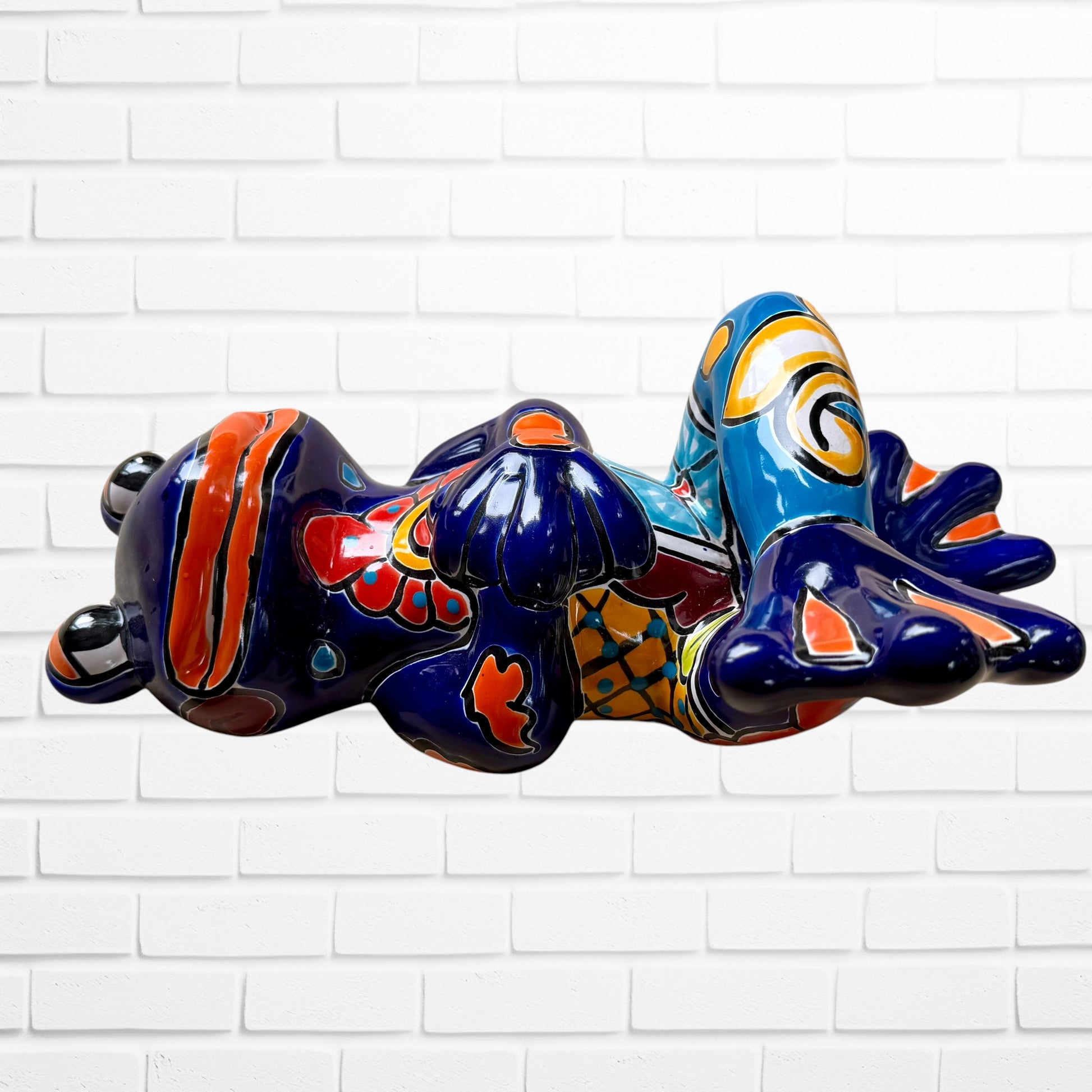 A colorful ceramic frog sculpture sitting cross-legged against a white brick wall background. The frog features a glossy deep blue body with vibrant orange, red, yellow, and light blue painted patterns and details right side view.