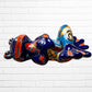 A colorful ceramic frog sculpture sitting cross-legged against a white brick wall background. The frog features a glossy deep blue body with vibrant orange, red, yellow, and light blue painted patterns and details right side view.