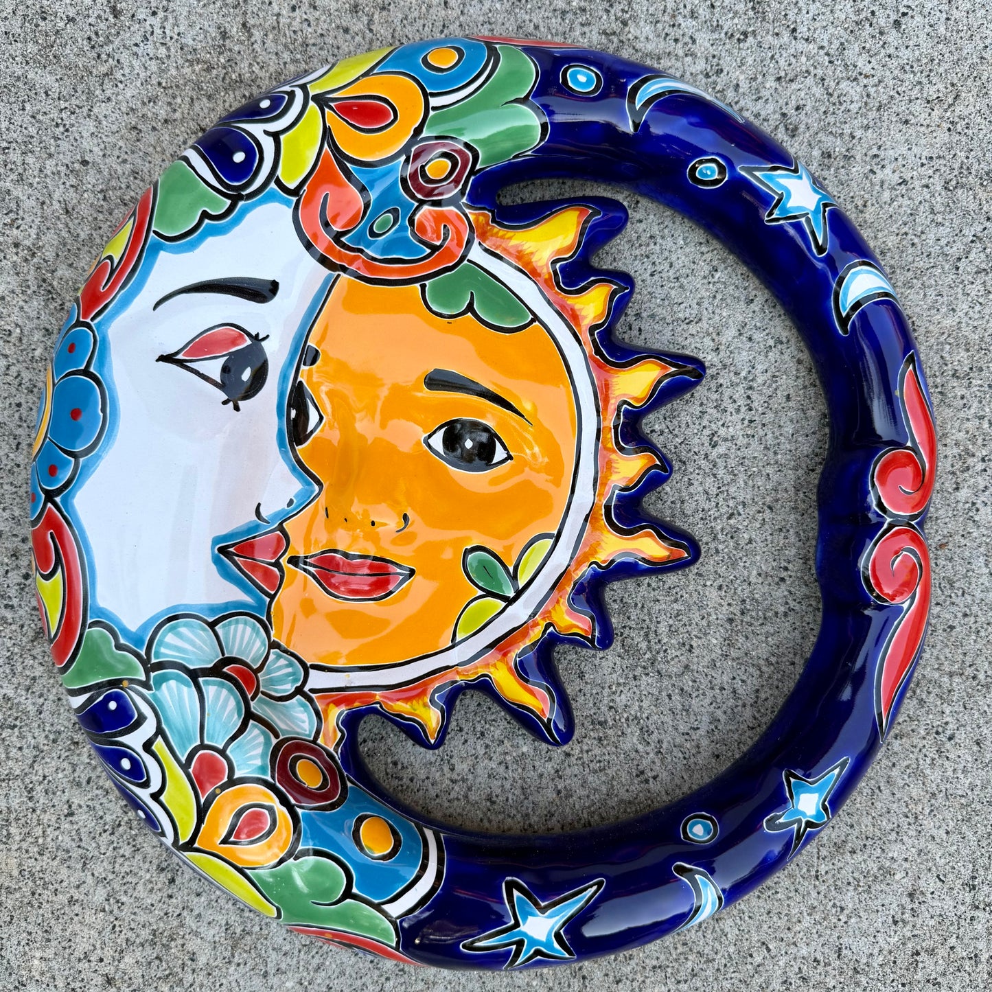 A colorful talavera wall hanging plaque featuring a stylized sun and moon face with decorative floral and star patterns, photographed from above on a gray stone surface.