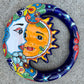 A colorful talavera wall hanging plaque featuring a stylized sun and moon face with decorative floral and star patterns, photographed from above on a gray stone surface.