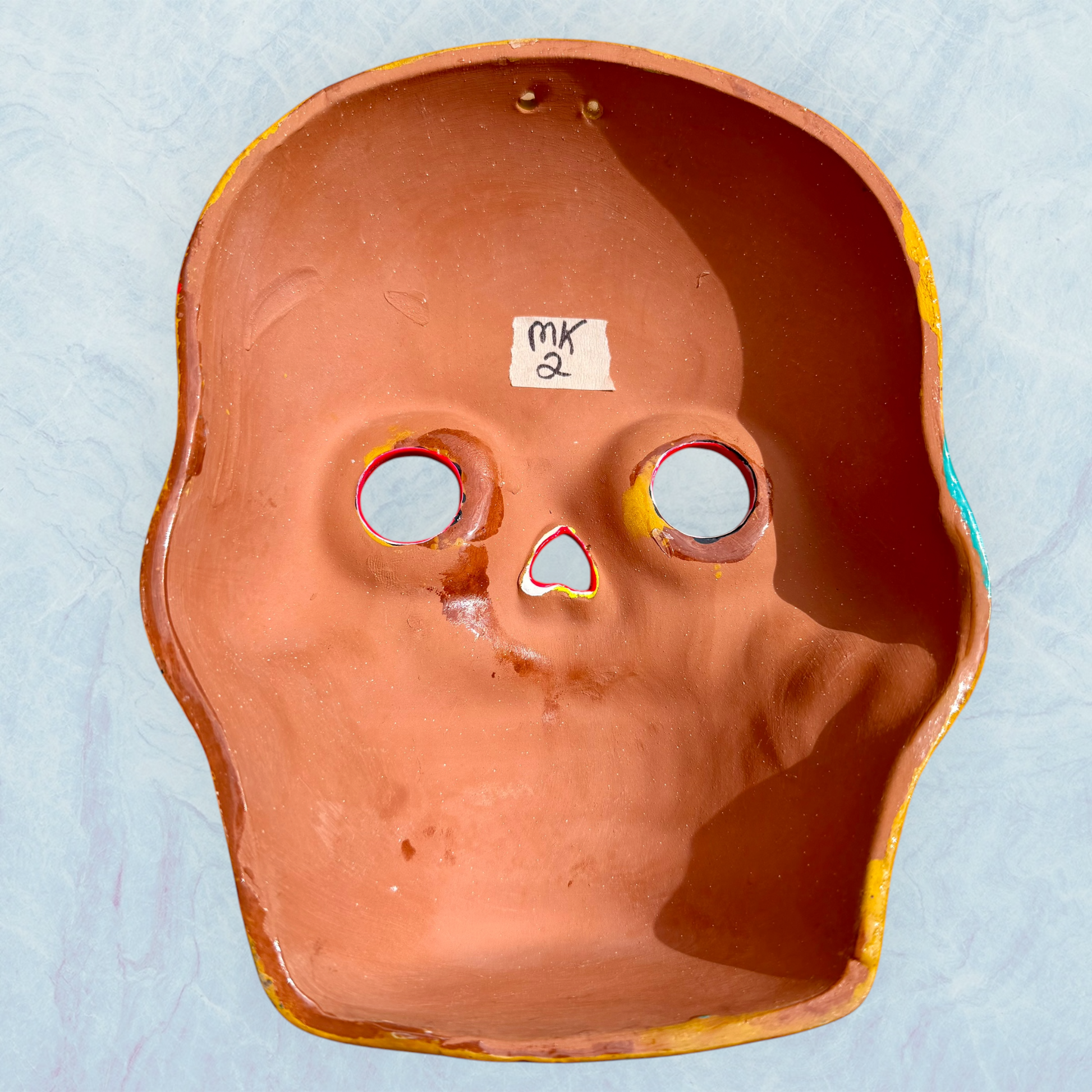 A clay, yellow with floral designs, Mexican-style skeleton skull face mask hanging on a soft blue surface, back.