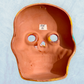 A clay, yellow with floral designs, Mexican-style skeleton skull face mask hanging on a soft blue surface, back.