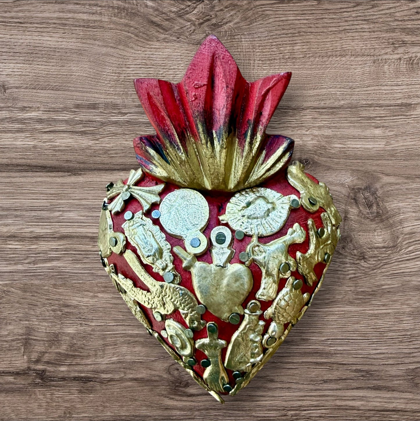 A decorative Milagros Sacred Heart featuring a red and gold painted heart adorned with charms, set against a rustic wood background.