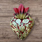 A decorative Milagros Sacred Heart featuring a red and gold painted heart adorned with charms, set against a rustic wood background.