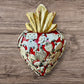 A Mexican Milagros heart in  red with a gold flaming top, covered in silver charms. 