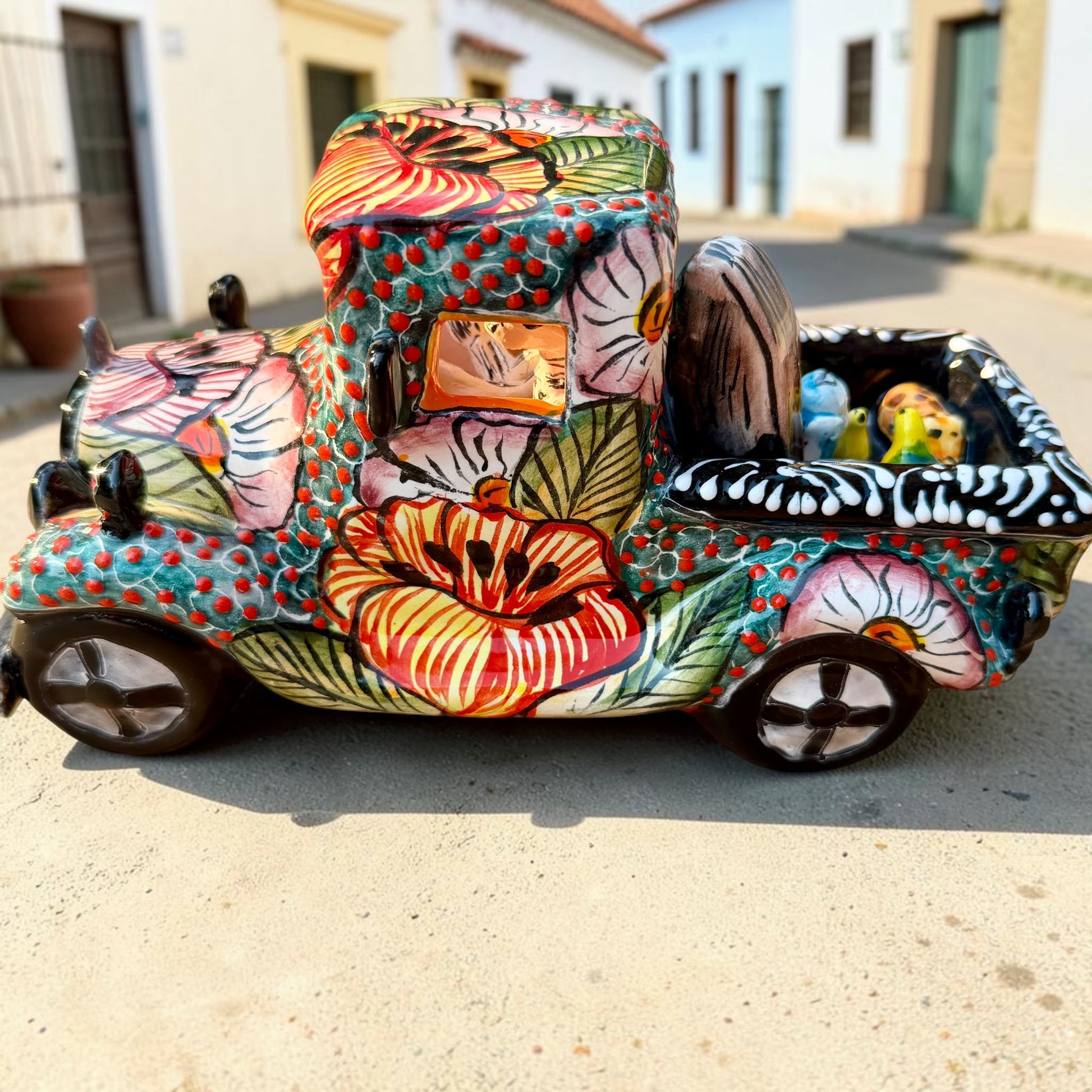 A decorative Talavera day of the dead pick up truck figurine two skeleton passengers. The truck is vibrant, with a hand-painted tropical floral pattern, photographed outdoors on a stone-paved street. This is the side of the pick up truck.
