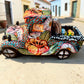 A decorative Talavera day of the dead pick up truck figurine two skeleton passengers. The truck is vibrant, with a hand-painted tropical floral pattern, photographed outdoors on a stone-paved street. This is the side of the pick up truck.
