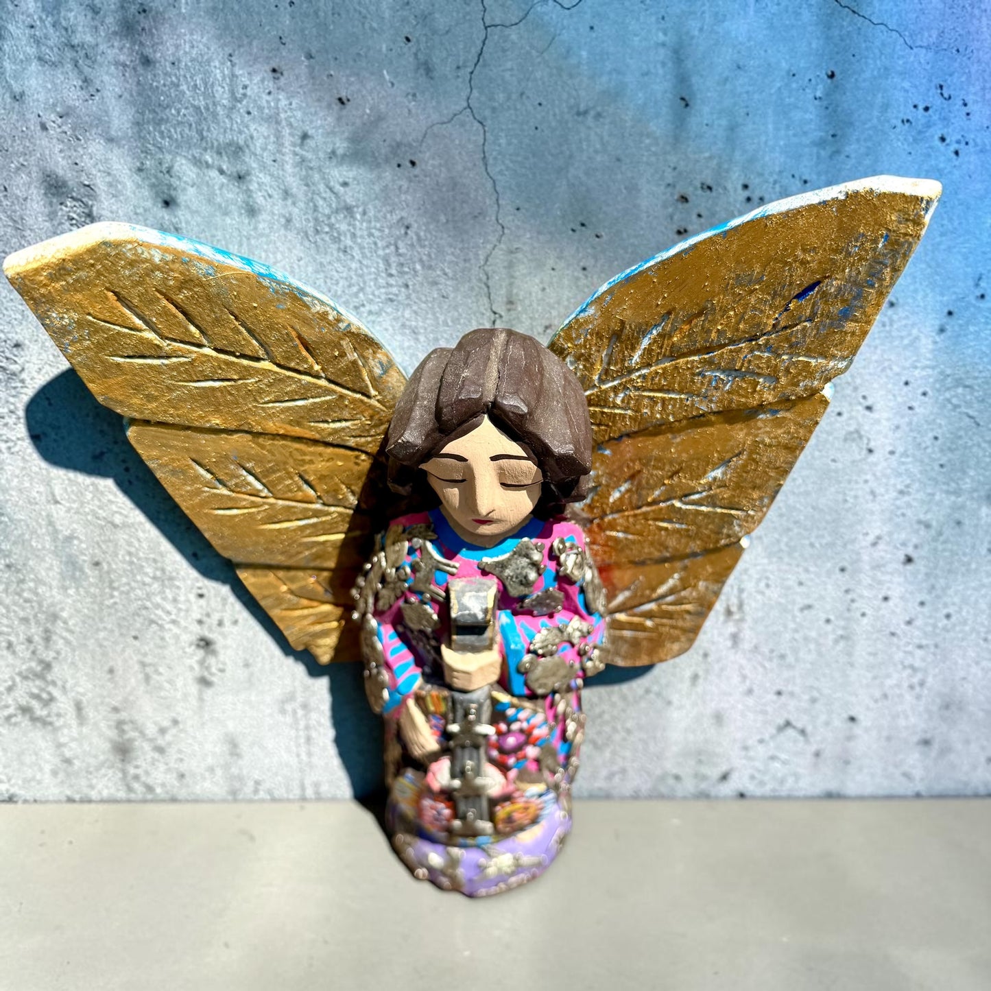 A handmade wood Milagros angel figurine with large gold wings, holding a guitar, dressed in a colorful patterned robe against a textured blue wall, top view.