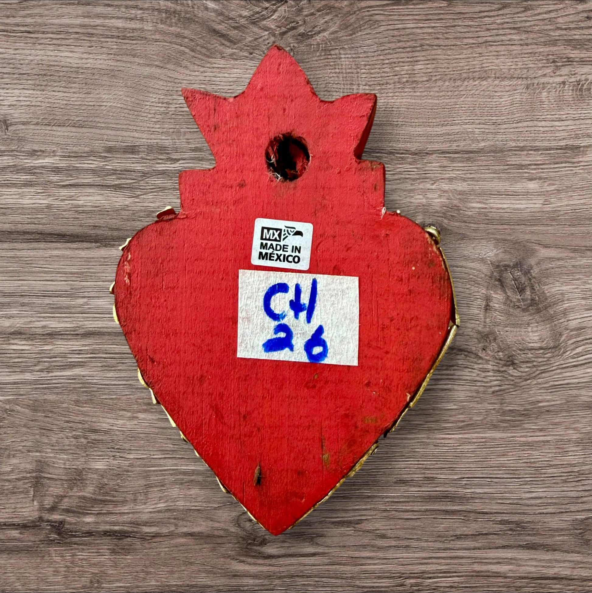 A decorative Milagros Sacred Heart featuring a red and gold painted heart adorned with charms, set against a rustic wood background, this is the back view.