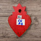 A decorative Milagros Sacred Heart featuring a red and gold painted heart adorned with charms, set against a rustic wood background, this is the back view.