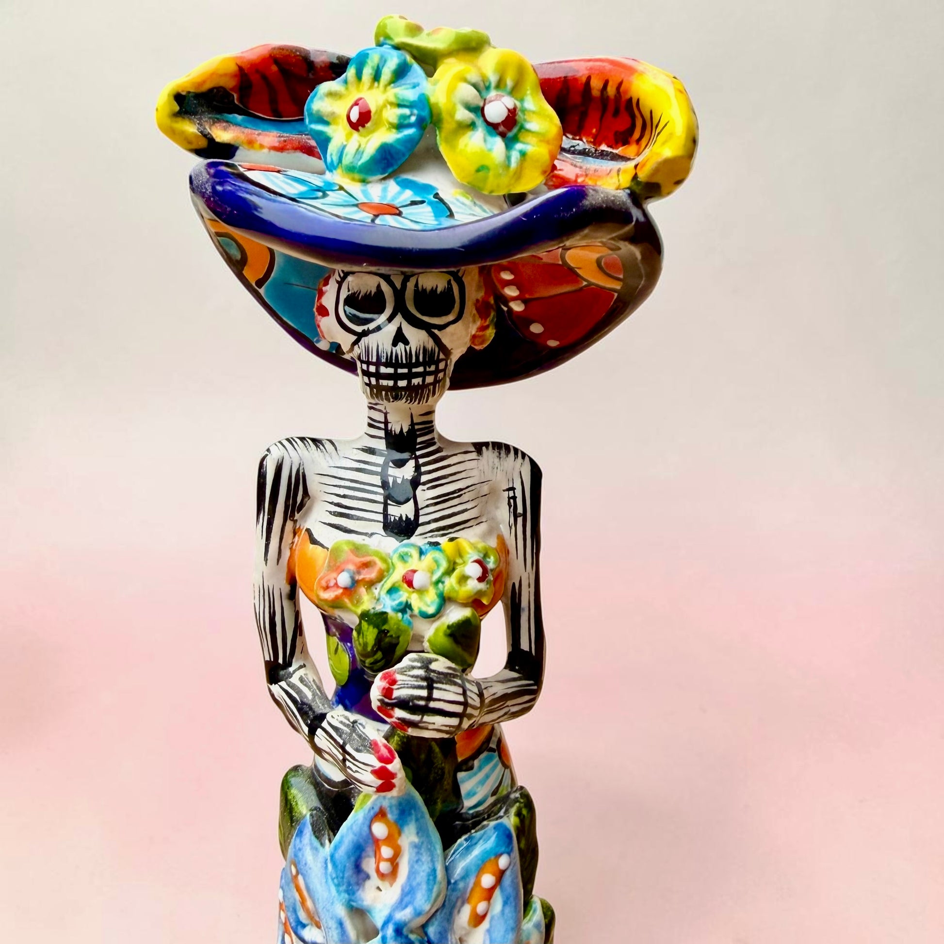 A Talavera Day of the Dead (Día de los Muertos) ceramic skeleton figurine with calla Lillies standing on a patterned base against a soft pink gradient background. This is a face close up.