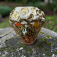 A Milagros religious figurine of Our Lady of Guadalupe with a child, standing on a mossy stone surface with a blurred outdoor garden background. She has a white halo and Lillies adorning the halo. Top view of halo.