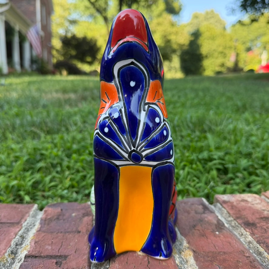 A colorful hand-painted Mexican Talavera Ceramic dog figurine sitting on a brick wall with a green grassy lawn and a house in the background front view.