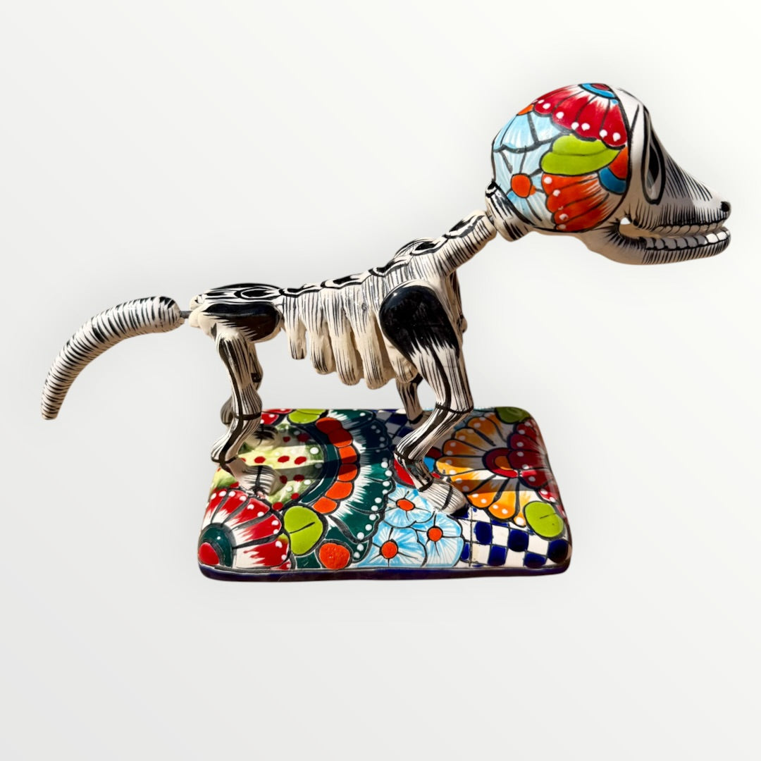 A ceramic Mexican-style skeleton dog figurine standing on a colorful floral base against a plain white background. Left side view of dog.
