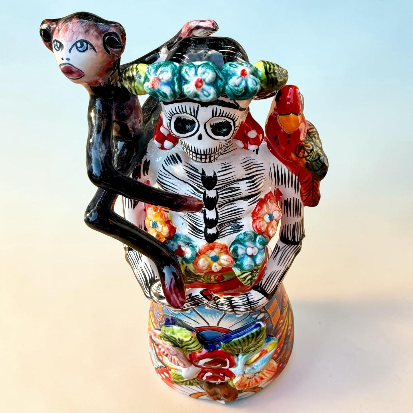 A Talavera Day of the Dead (Día de los Muertos) Frida figurine  with a monkey on her shoulder and a red parrot on her other shoulder, set against a light blue gradient background. View from top down, highlighting monkey and parrot.