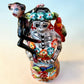 A Talavera Day of the Dead (Día de los Muertos) Frida figurine  with a monkey on her shoulder and a red parrot on her other shoulder, set against a light blue gradient background. View from top down, highlighting monkey and parrot.