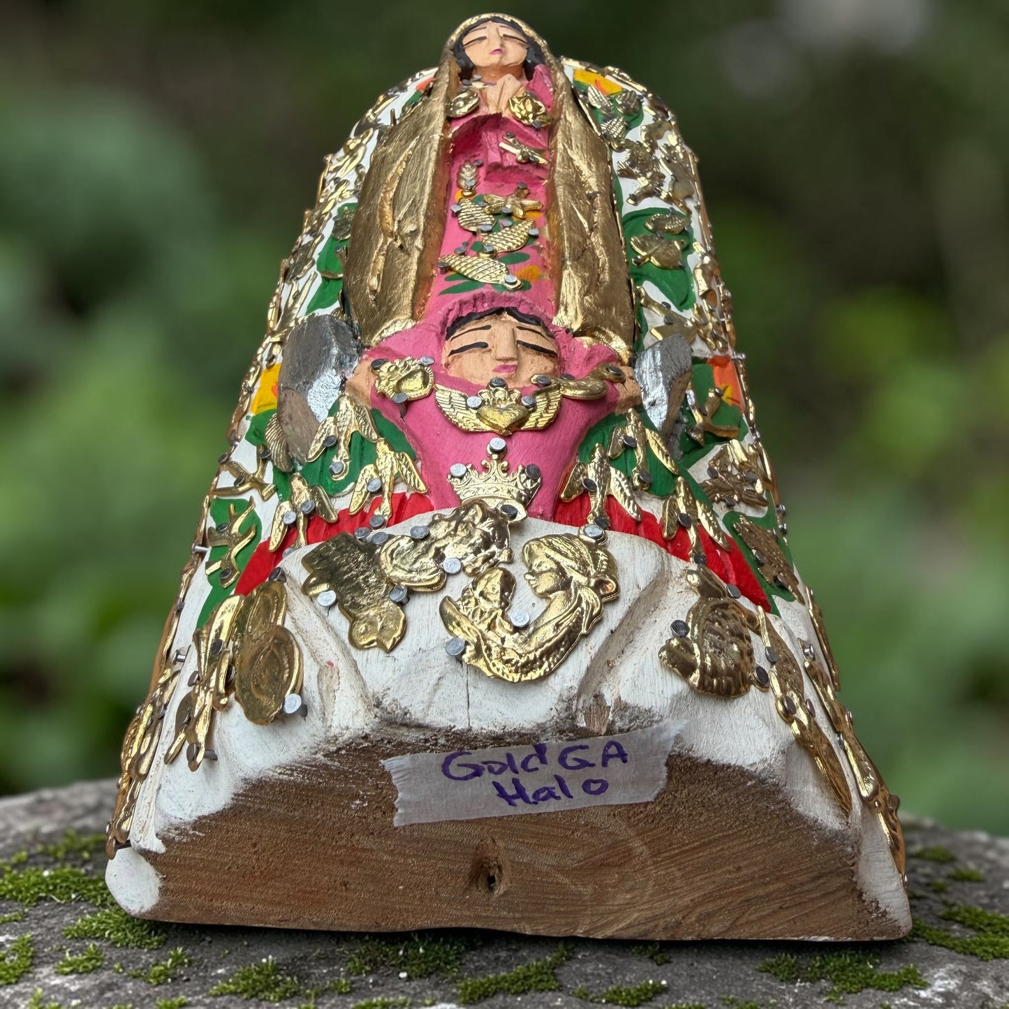 A Milagros religious figurine of Our Lady of Guadalupe with a child, standing on a mossy stone surface with a blurred outdoor garden background. She has a white halo and Lillies adorning the halo. Bottom view of figurine.