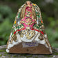 A Milagros religious figurine of Our Lady of Guadalupe with a child, standing on a mossy stone surface with a blurred outdoor garden background. She has a white halo and Lillies adorning the halo. Bottom view of figurine.