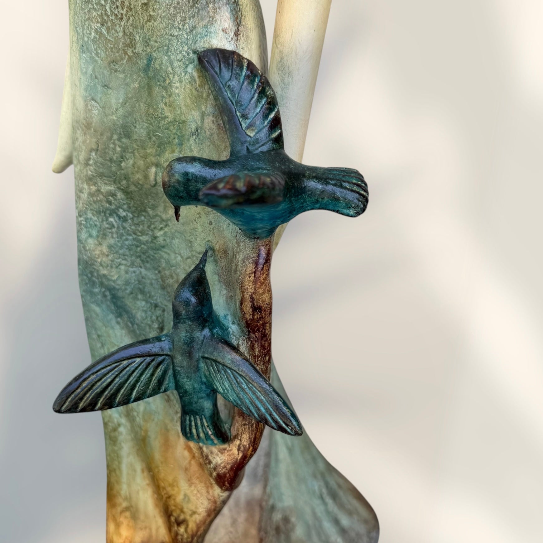 A Loretta Bertoli statue of a faceless woman in a long flowing dress with two hummingbirds, displayed against a stacked stone tile wall on a light tiled floor. This is a close-up of the hummingbirds.