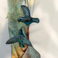 A Loretta Bertoli statue of a faceless woman in a long flowing dress with two hummingbirds, displayed against a stacked stone tile wall on a light tiled floor. This is a close-up of the hummingbirds.