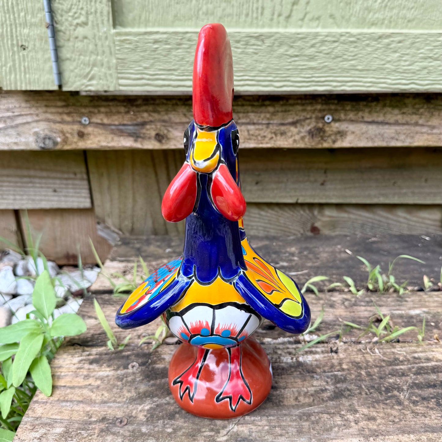 A colorful hand-painted ceramic rooster figurine standing on a rustic wooden surface in front of a green wooden door, front view.
