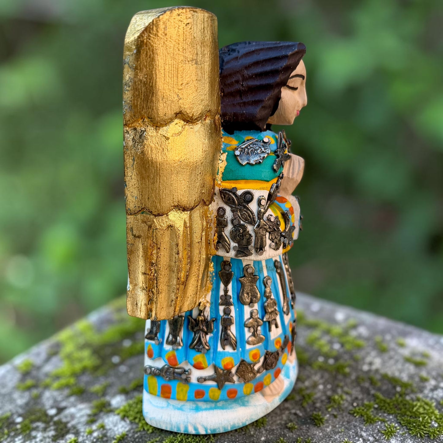 A Milagros guardian angel figurine with gold wings, dark hair, and a blue, white, and gold embroidered dress, posed outdoors on a stone surface with a blurred green background. thesis there left side view.