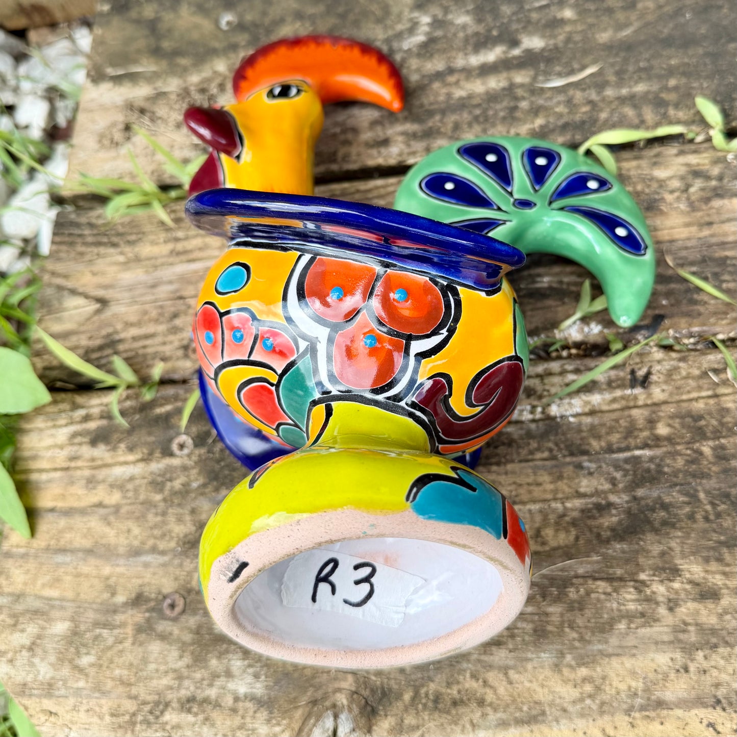 A colorful hand-painted ceramic Mexican-style rooster figurine standing on a rustic wooden surface outdoors, rooster bottom sculpture view.