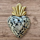 A Milagros heart featuring a gold textured top and a black base covered in raised silver-toned charms displayed on a wooden wall.