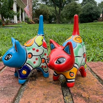 Two hand-painted Mexican-style ceramic cat figurines, one blue and one red, decorated with colorful geometric patterns, standing on a brick surface outdoors right side view