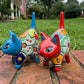 Two hand-painted Mexican-style ceramic cat figurines, one blue and one red, decorated with colorful geometric patterns, standing on a brick surface outdoors right side view