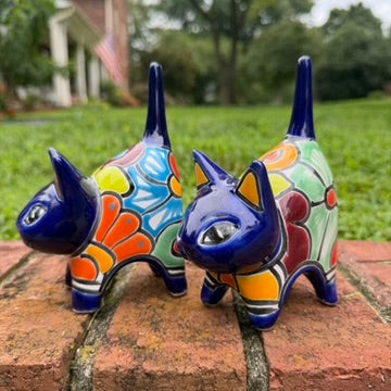 Two colorful hand-painted talavera ceramic cat figurines in cobalt blue with multi color designs standing on a brick path with a green lawn right side view.