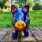 A glossy talavera pig figurine decorated with vibrant hand-painted patterns in blue, yellow, red, green, and white, standing on a brick surface with a blurred outdoor garden background, front face closeup. 