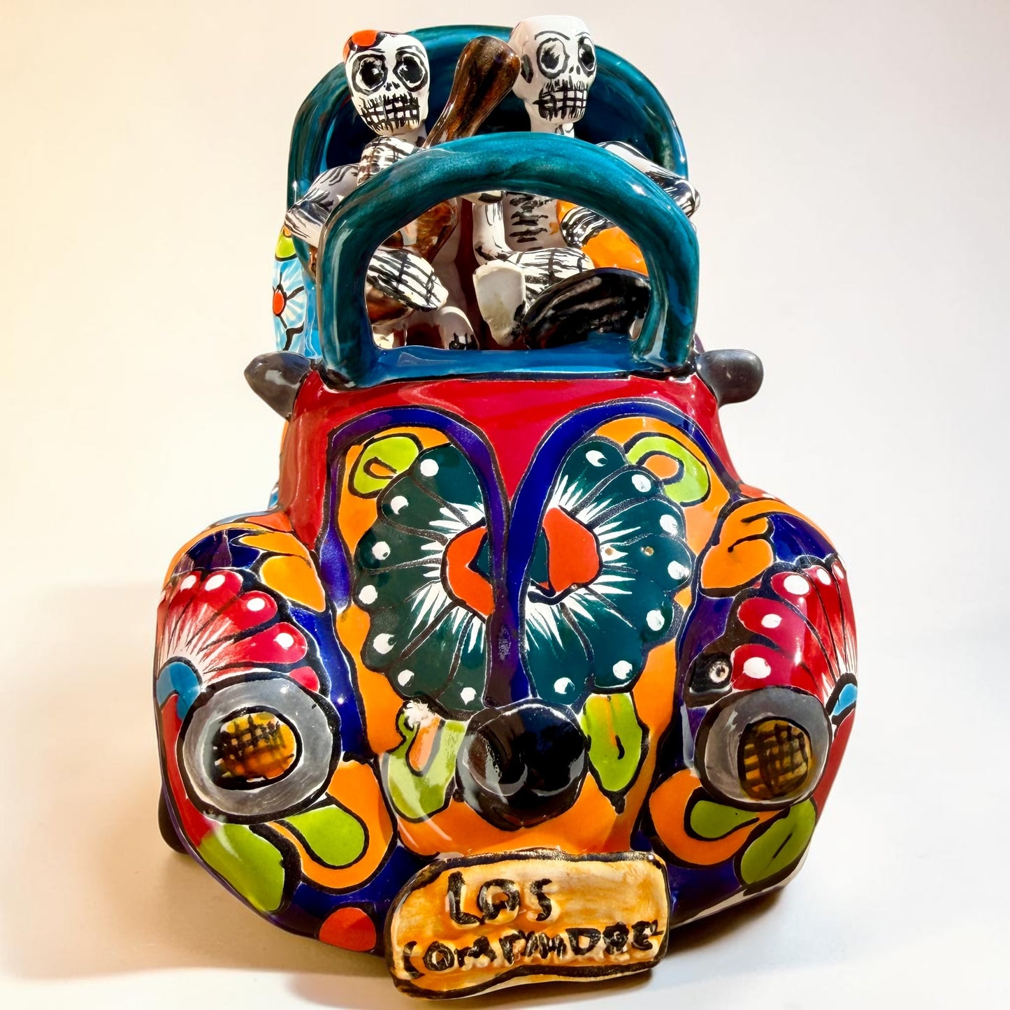 A colorful hand-painted ceramic Mexican-style convertible car figurine with Day of the Dead skeleton characters inside, shown on a neutral beige background. Front view of convertible car with license tag.