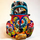A colorful hand-painted ceramic Mexican-style convertible car figurine with Day of the Dead skeleton characters inside, shown on a neutral beige background. Front view of convertible car with license tag.