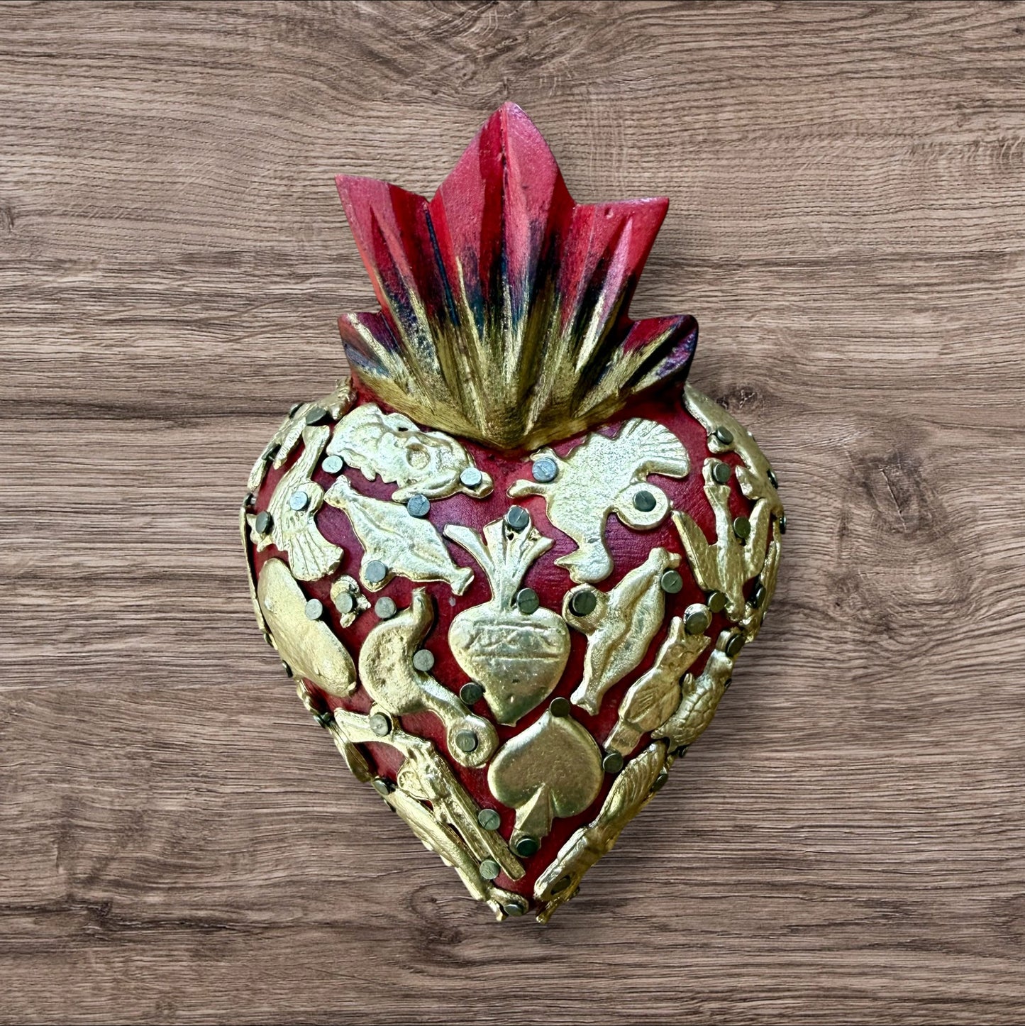 A Mexican Milagros heart in red with a gold and red flaming top, covered in antiqued charms. 