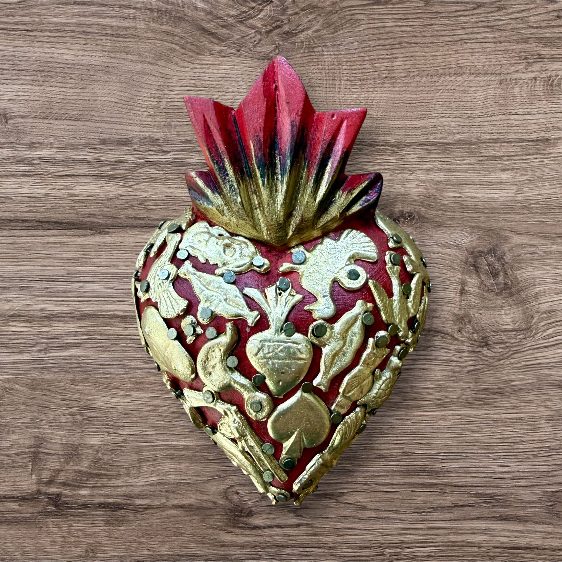 A Mexican Milagros heart in red with a gold and red flaming top, covered in antiqued charms. 