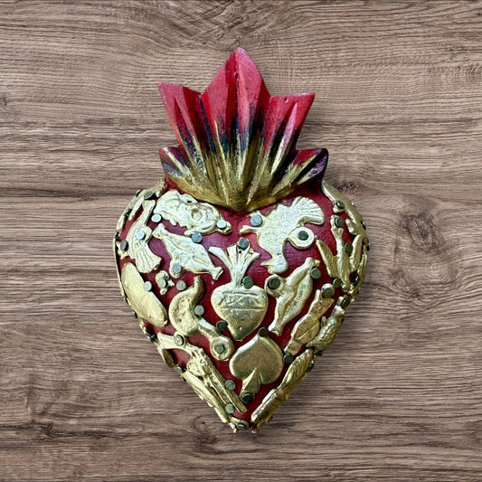 A Mexican Milagros heart in red with a gold and red flaming top, covered in antiqued charms. 