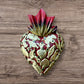 A Mexican Milagros heart in red with a gold and red flaming top, covered in antiqued charms. 