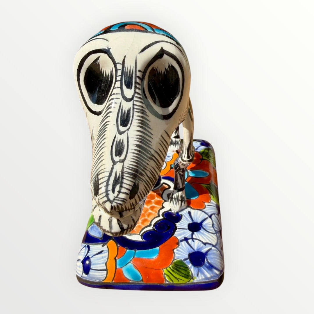 A ceramic Mexican-style skeleton dog figurine standing on a colorful floral base against a plain white background. Front view of dog showing face.