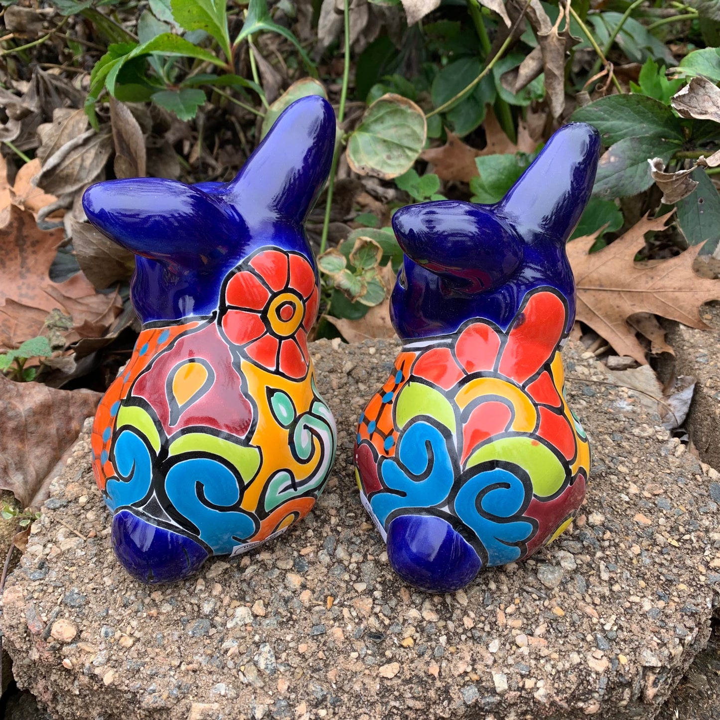 Two colorful Mexican Talavera rabbit figurines on a stone surface with greenery in the background set of two back view