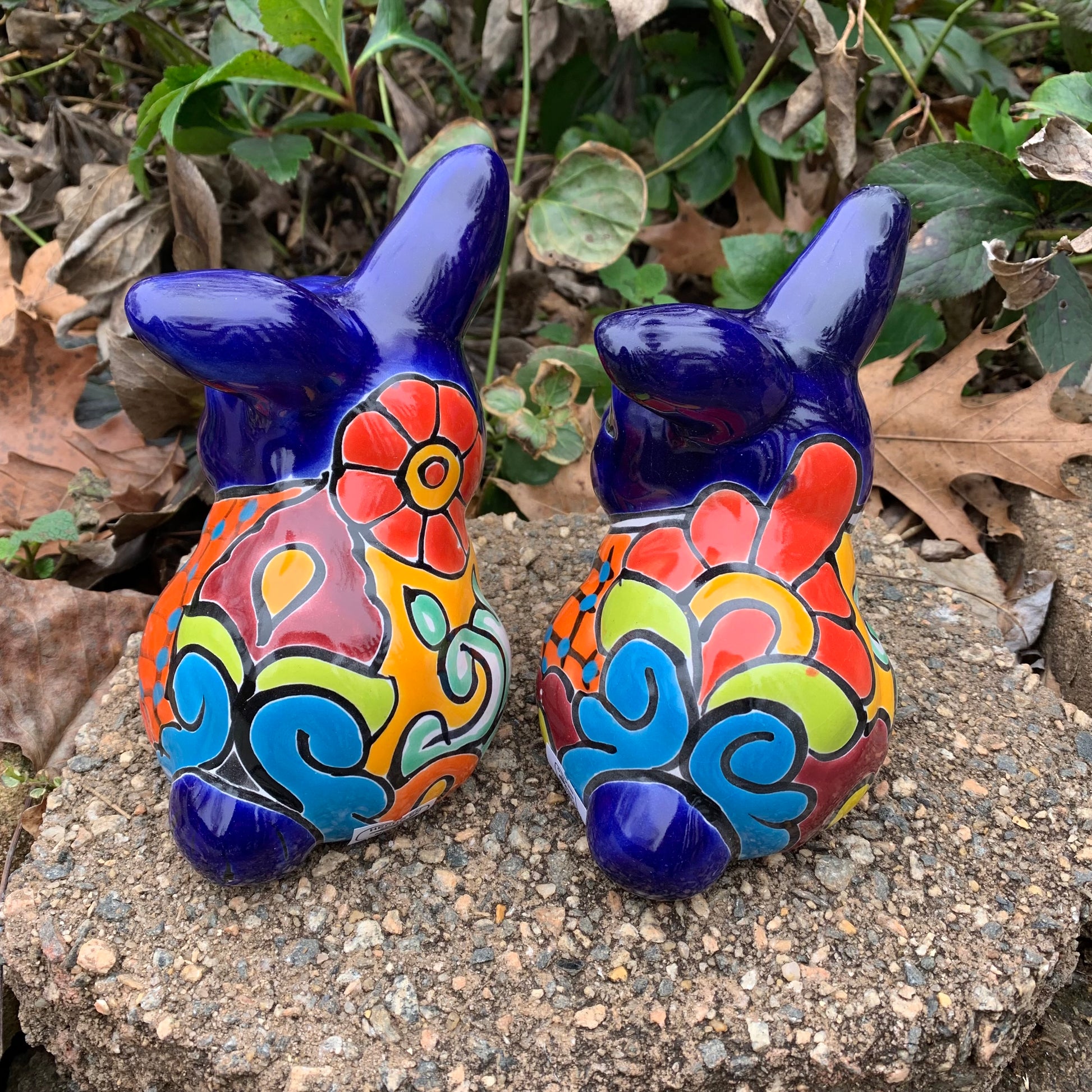 Two colorful Mexican Talavera rabbit figurines on a stone surface with greenery in the background set of two back view