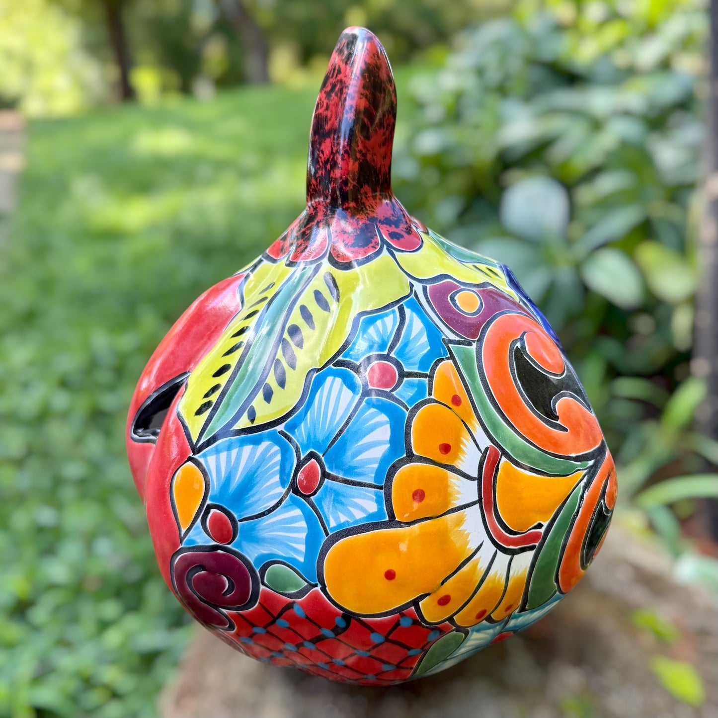 A hand-painted ceramic jack-o'-lantern pumpkin with a red face, black carved eyes and mouth, and a multicolored floral pattern on a garden background. Right side view of pumpkin.