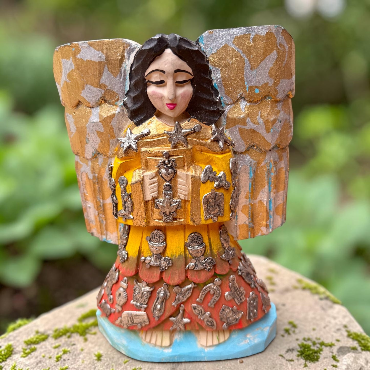 A handcrafted wooden Milagros angel figurine with large wings, decorated with metallic silver charms and painted in yellow, and orange, standing on a mossy stone with a blurred green garden background.