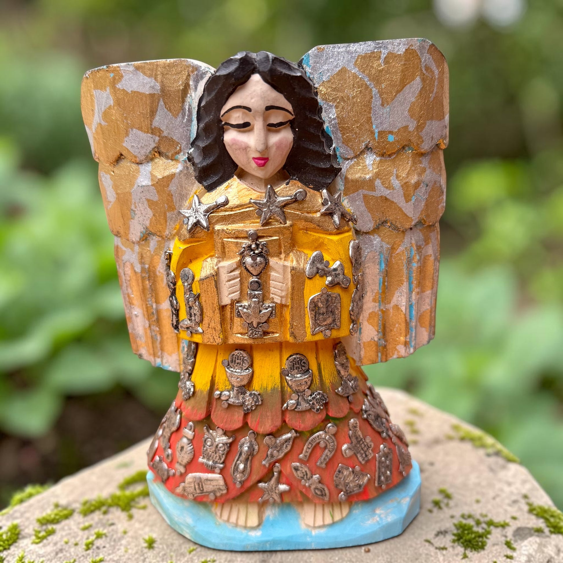 A handcrafted wooden Milagros angel figurine with large wings, decorated with metallic silver charms and painted in yellow, and orange, standing on a mossy stone with a blurred green garden background.