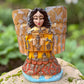 A handcrafted wooden Milagros angel figurine with large wings, decorated with metallic silver charms and painted in yellow, and orange, standing on a mossy stone with a blurred green garden background.