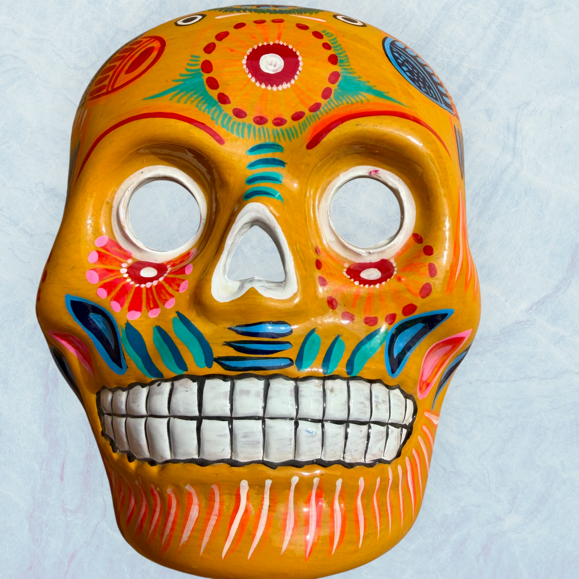 A clay,  golden yellow with floral designs, Mexican-style skeleton skull face mask hanging on a soft blue surface.