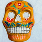 A clay,  golden yellow with floral designs, Mexican-style skeleton skull face mask hanging on a soft blue surface.