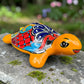 A glossy ceramic turtle figurine with a bright yellow body and a highly decorated shell in red, blue, yellow, black, and white, resting on a stone surface with a blurred garden background.